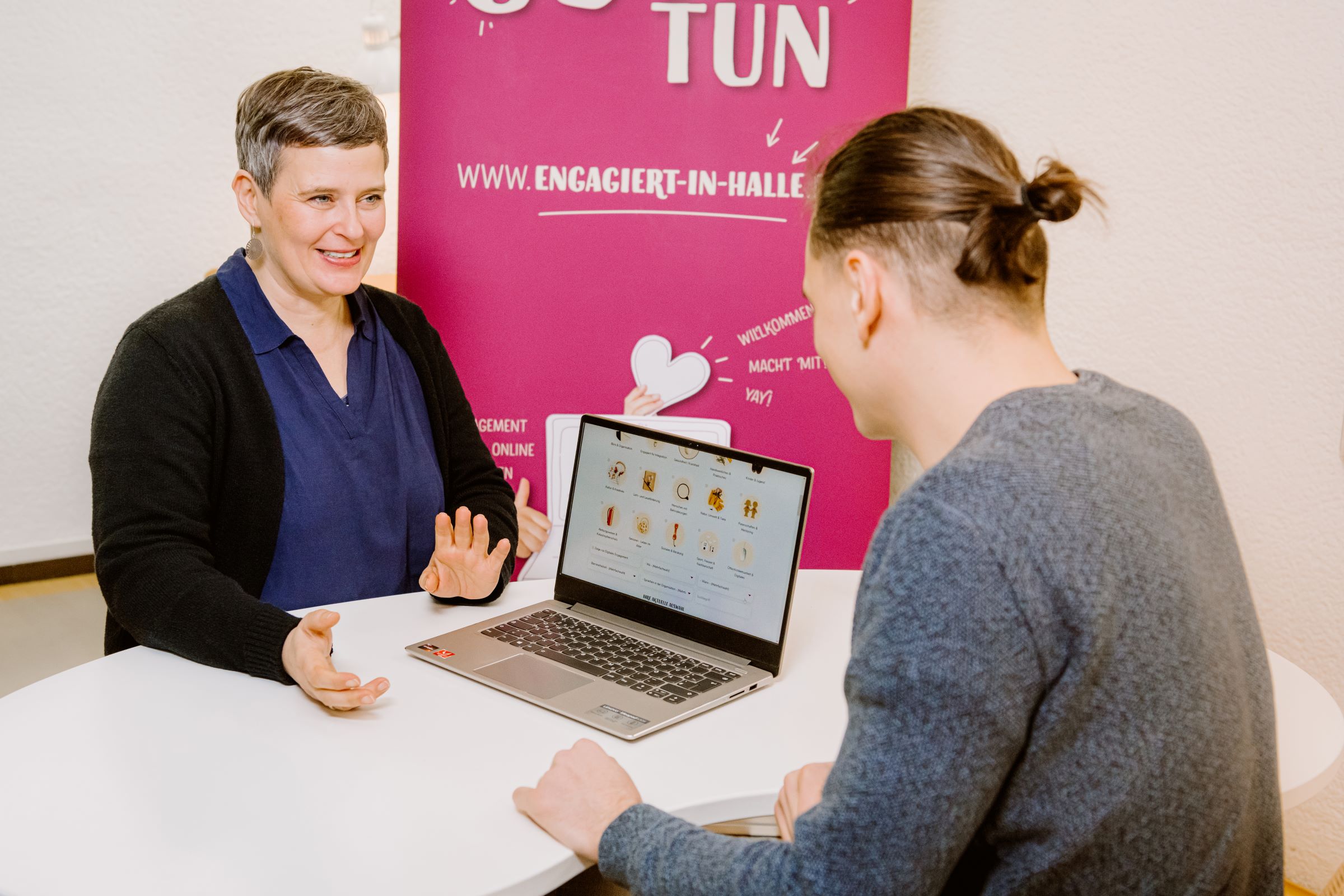 Eine Frau und ein Mann sitzen sichgegenüber an einem Tisch, die Frau zeigt auf einen Laptop