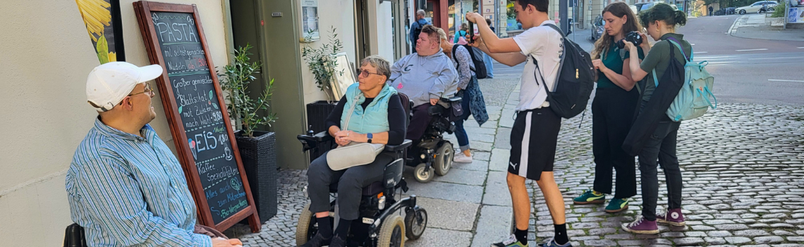 Eine Gruppe aus Fußgänger:innen und Rollstuhlfahrer:innen steht vor einem Restaurant in Halle, einige haben ein Handy oder eine Kamera in der Hand. Sie sind Teilnehmende einer Wheelmap-Aktion.