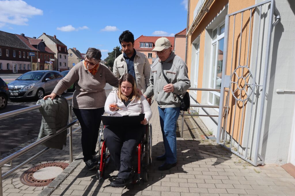 Eine Gruppe bei einer Wheelmap-Aktion steht vor einer Einrichtung, zwei Männer und zwei Frauen, eine davon im Rollstuhl