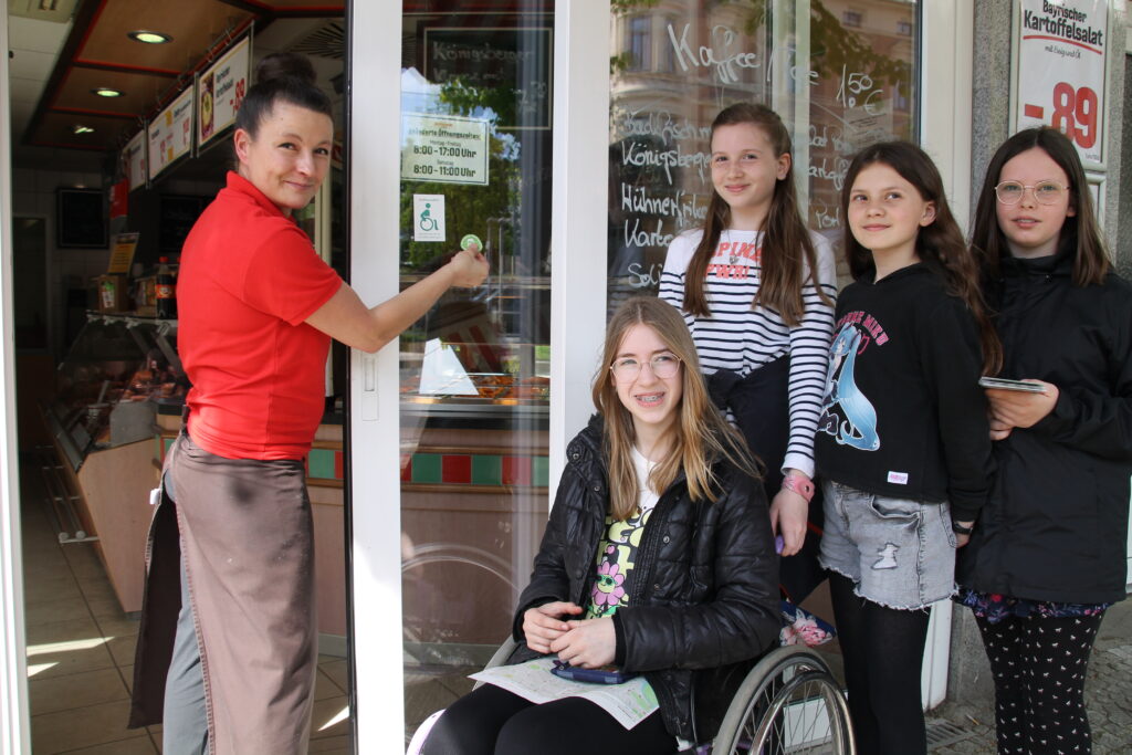 Links steht eine Verkäuferin, die einen Wheelmap-Aufkleber an die Ladentür klebt, daneben 4 Schülerinnen, eine davon im Rollstuhl