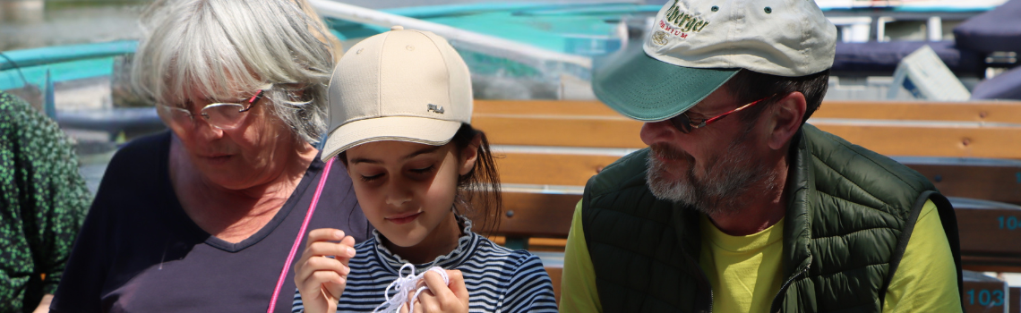Ein Kind in der Mitte schaut auf einen Wollfaden, links eine Frau; der Mann rechts schaut liebevoll zum Kind